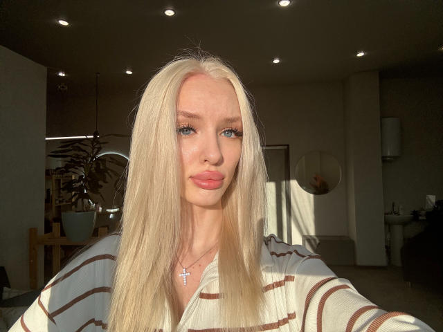 LexyGlam has long, straight blonde hair, blue eyes, and full pink lips. She's wearing a striped shirt, with a cross necklace, and sunlight highlights her face.