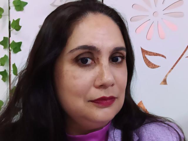 BabsBloom ha i capelli castano scuro, la pelle chiara, una piccola macchia sul viso e indossa un rossetto rosa con una maglietta viola. La sua espressione è neutra..