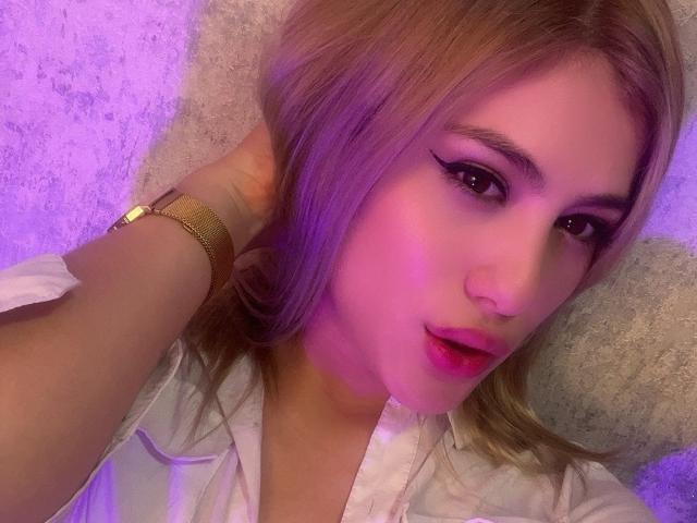 AdelaLaurent with pinkish-purple hair, thick black eyeliner, and pink lipstick, wearing a white shirt, gold wristwatch, and pinkish-purple light on her face. Her hand's in her hair.