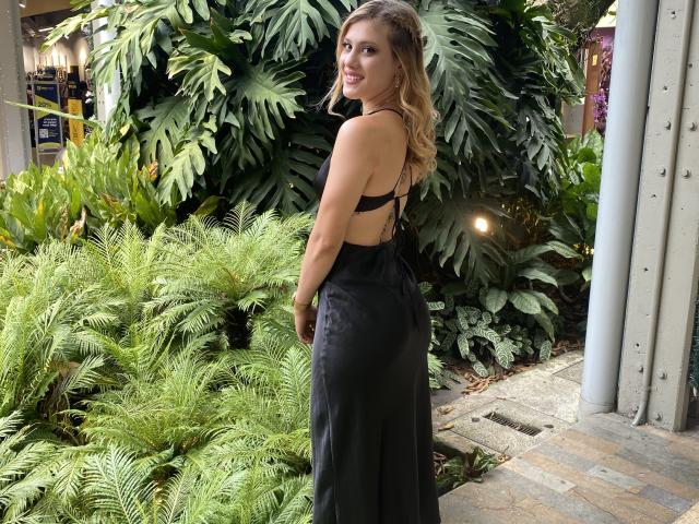 Adela Laurent mit langen blonden Haaren, lächelnd, trägt ein schwarzes, trägerloses Kleid, das ihr Gesäß zeigt, und steht vor üppigen, grünen Pflanzen. Ihre Brüste sind klein, und sie ist süß..