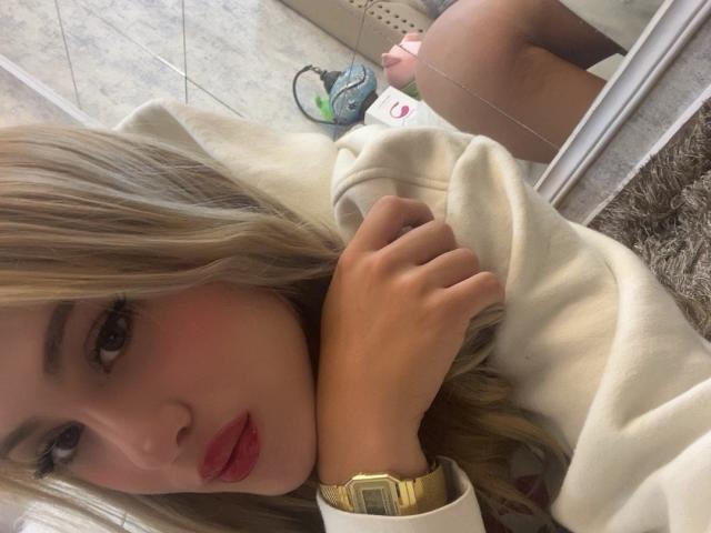 Adela Laurent mit blonden Haaren, roten Lippen und einer goldenen Uhr, hält ihre weiße Kapuzenjacke und betrachtet einen Spiegel, der ihr rundes Gesäß zeigt. Ihre Hand liegt auf ihrer Brust..