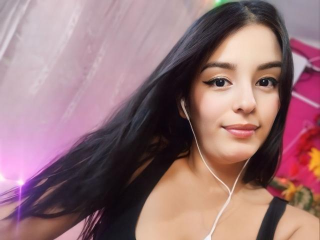 TalianaSquirt has long black hair, wearing a black tank top, white earbuds, and a slight smile. Her tits are small and her skin is light. She's cute.
