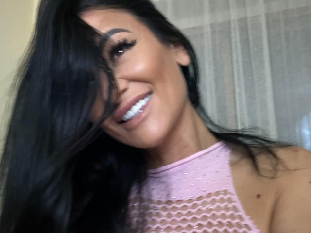 AndraD has long black hair covering one eye, smiling with white teeth, wearing a pink mesh top, showing her tan skin and small moles on her neck.