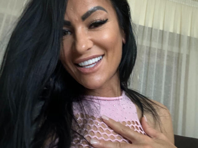 AndraD has black hair, smiling with white teeth, wearing a pink fishnet top, exposing her tits, and holding her right breast with her hand.