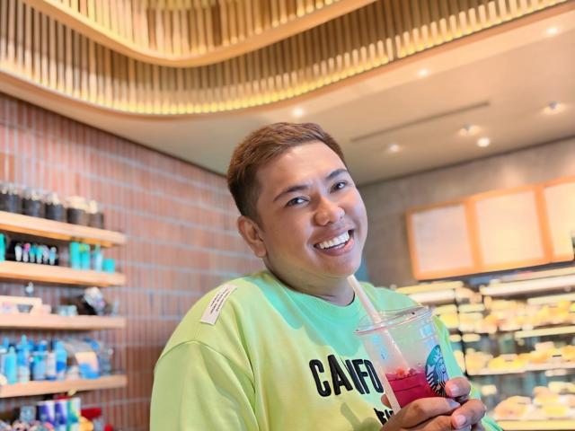 OneSpiceDiva with short brown hair, smiling, wearing a green shirt, holding a clear plastic cup with a pink drink and blue straw, in a brightly lit cafe.