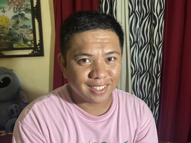 OneSpiceDiva, chubby Asian dude with short black hair, wearing a pink shirt, smiling with shiny skin, sitting in front of red and zebra-striped curtains. Stuffed blue creature on left.
