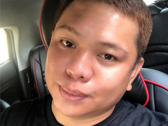 OneSpiceDiva's face, brown skin, short brown hair, brown eyes, slight smile, black shirt, black and red car seat, light from left, visible pores, slight nose bump.
