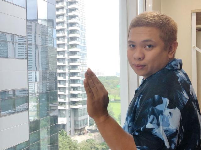 OneSpiceDiva with short blond hair, blue and black patterned shirt, standing by window, right hand up, looking over shoulder, cityscape with tall buildings in background, green park below.