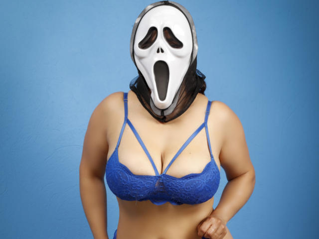 Becck with a white Ghost mask, blue lace bra, and visible boobs, wearing a black sheer scarf, against a blue background. Her hands are on her hips.