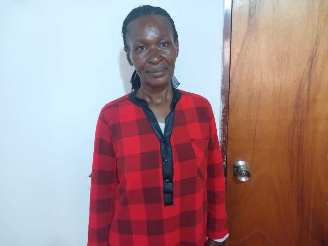 HotMatureSexy69, dark-skinned, medium-sized tits, wearing red and black checkered shirt, black collar, standing in front of wooden door, looking at camera with slight smile.