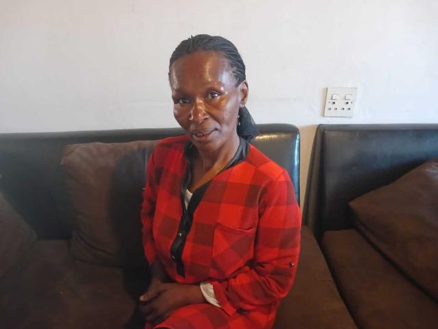 HotMatureSexy69 with dark skin, braided hair, wearing a red checkered shirt, sitting on a brown leather couch, looking at the camera with a slight smile.