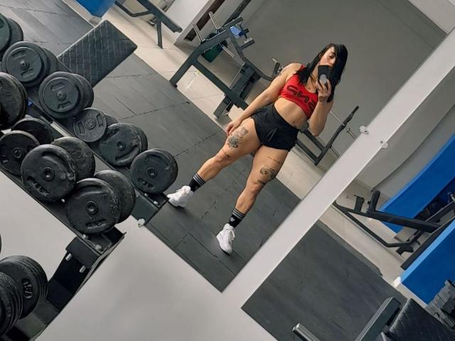 YeiSela with tanned skin, black hair, red sports bra, black shorts, white socks, white sneakers, visible tattoos on thighs, standing in gym, mirror selfie, weights in foreground.