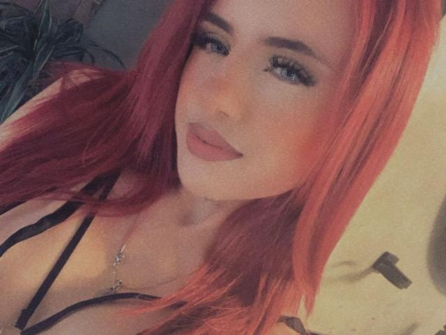 Nia Spencer mit roten Haaren, blauen Augen und einem leichten Lächeln, trägt ein schwarzes Top mit dünnen Trägern, das ihre Brüste zeigt, und eine silberne Halskette. Ihre Lippen sind leicht geöffnet..