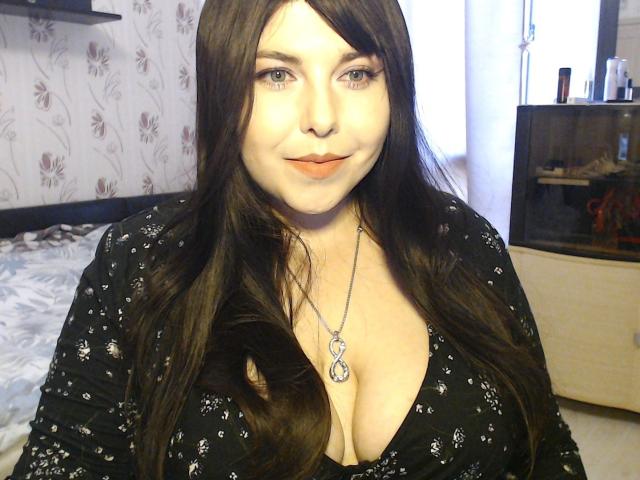 WhitteLotus has long brown hair, blue eyes, and a fair skin tone. She's wearing a black patterned top with a low neckline, showing her large boobs. She's smiling slightly.