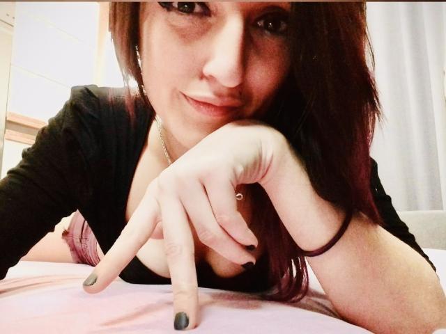 MissCherryOnTop, avec ses cheveux foncés, son vernis à ongles noir et son haut noir, appuyée sur un lit rose, pointant du doigt, souriante, portant une bague et un bracelet..