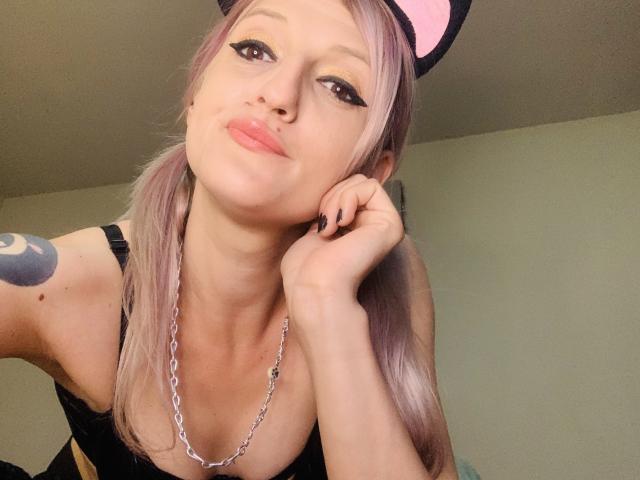 MissCherryOnTop with pink hair, cat ears, and pink nose, looking down, black lingerie, silver chain, tattoo on right arm, black nail polish, and a cute, sexy smile.