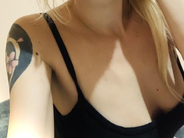 MissCherryOnTop's bare shoulders, black strap top, and visible tattoo of a pink flower on her upper arm, with blonde hair falling to the side, showing her tits.