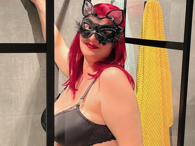 MelanieFrancaiseDuo: una donna con capelli rossi, una maschera di pizzo nero e un reggiseno nero, che mostra il seno, in piedi in un bagno con asciugamani gialli e verdi sullo sfondo..