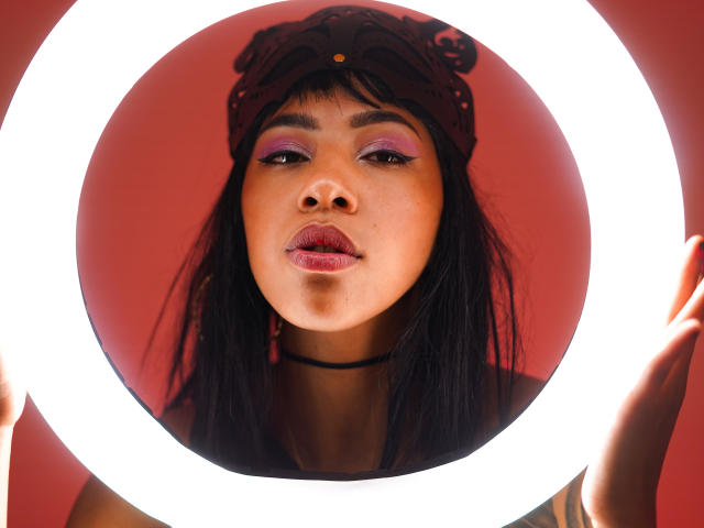 InannaIsshtar with dark hair, purple eyeshadow, black choker, and headband, holding a bright ring light, showing her face and pink lips, against a red background.