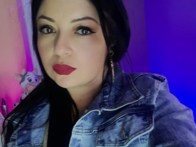 NatalyHami, mit dunklem Haar, rotem Lippenstift und einer blauen Jeansjacke, trägt goldene Ohrringe, hat ein attraktives Gesicht und blickt mit einem leichten Lächeln in die Kamera..