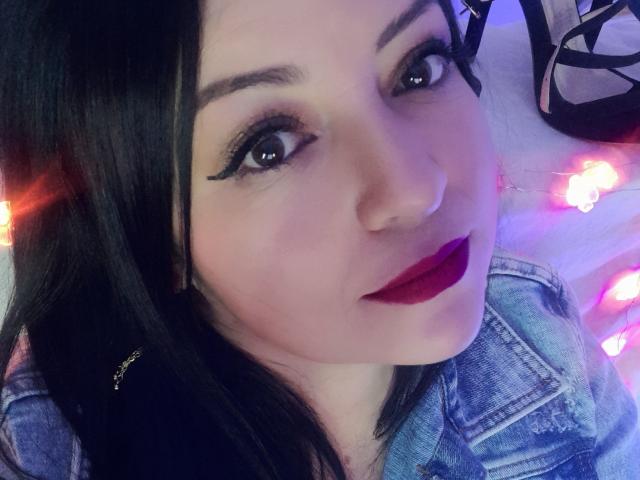 NatalyHami's face, with dark hair, thick black eyeliner, and deep red lipstick, wearing a blue denim jacket, has a cute smile, and her eyes are looking at the camera.