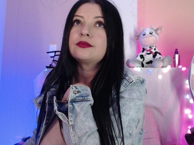 NatalyHami with long black hair, red lipstick, and a denim jacket, partially exposing her tits, stares up with a subtle smile. A stuffed cow is on the right.