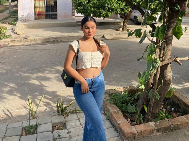 MiiaPalmer, avec de petites seins, portant un crop top blanc, un jean bleu évasé, un sac noir et des couettes, debout près d'un arbre, regardant vers le bas, dans un cadre extérieur ensoleillé..