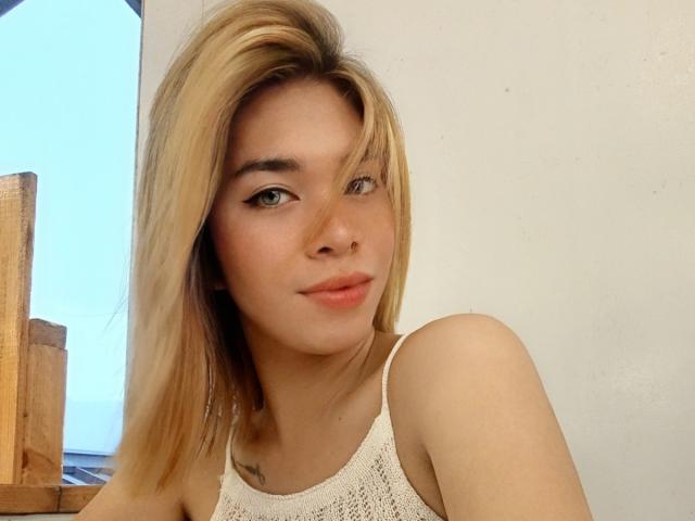 Godin Hera met blond haar, groene ogen en een neusring, gekleed in een wit, gebreid tanktopje, waarbij haar borsten zichtbaar zijn, met een lichte glimlach, in een heldere, binnenruimte..