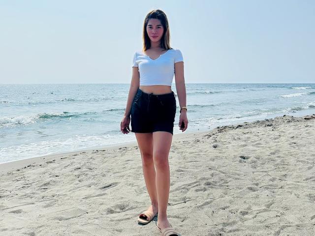 GoddesHera staat op een strand met haar kleine borsten, gekleed in een witte crop top en een zwarte rok, waarbij haar blote billen te zien zijn in een zwarte string, en bruine sandalen..