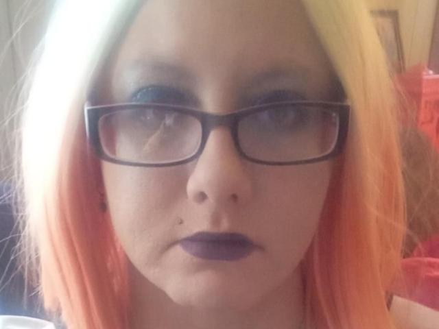 StarLove69 ha la pelle chiara, rossetto viola, occhiali neri e capelli con sfumatura arancione-gialla. Il suo viso è al centro dell'immagine e ha un'espressione seria..