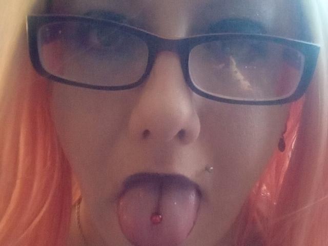 Het gezicht van StarLove69, met zwarte brillen, roze haar, een neusring en haar tong uit, met een tongpiercing. Haar tong is doorboord met een rode bal. Haar ogen zijn lichtjes samengeknepen..
