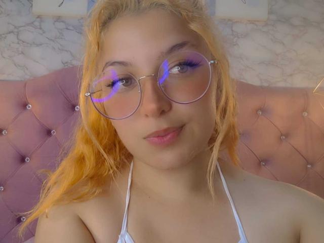AmaliaRosse with orange hair, wearing round glasses with purple lenses, white halter top, sitting on pink tufted headboard, smiling softly, showing small tits and cute face.
