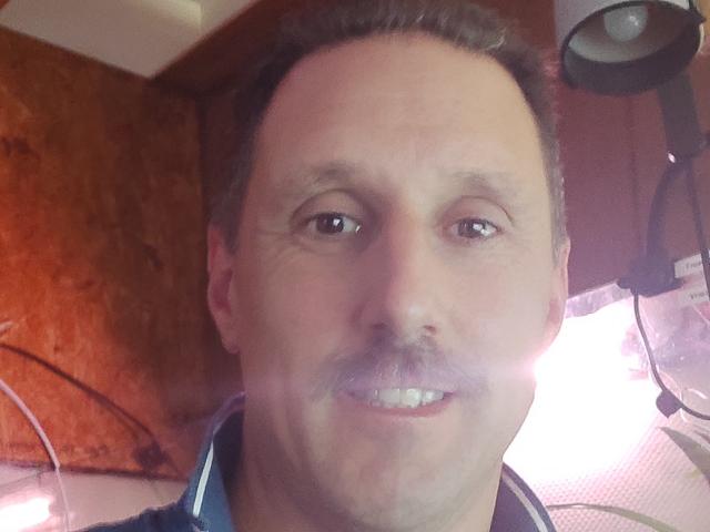 JefHotMan hat einen glatten Kopf, braune Augen und einen kleinen Schnurrbart mit einem leichten Lächeln. Er trägt ein blaues Hemd, und im Bild ist ein leichter Schein zu sehen..