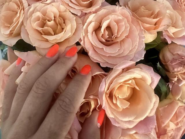 Stefanni's hand with neon orange nails rests on a bunch of pink, rose-like flowers, showing her fingers and colorful nails against the soft, petal-filled background.