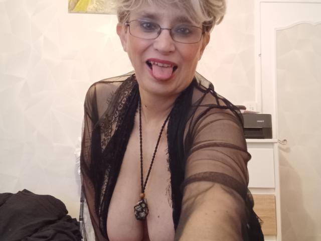 Samantha, una donna matura con i capelli biondi corti, occhiali e un abito nero trasparente, che mostra la lingua, con un seno prosperoso e una collana, in un selfie..