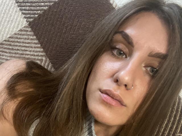 Kametia has long brown hair, green eyes, and a nose ring, with slightly parted pink lips, looking at the camera. She's lying on a striped brown and beige couch.