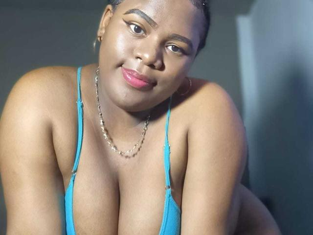EbonySkyX with shiny brown skin, blue bra, and big tits, leaning forward, smiling with pink lips, wearing a chain necklace, and hoop earrings, in a well-lit, slightly shadowed photo.