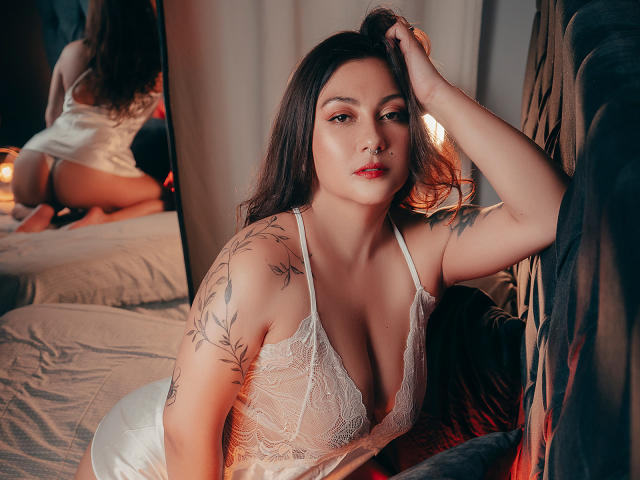 Karlaparadise with tits in a white lace lingerie, tattoos on her arm, brown hair, red lips, hand in hair, mirror reflecting her ass, candles in the background.