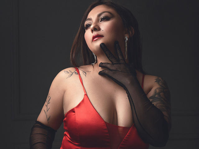 Karlaparadise with red satin dress, black gloves, tattoos, and makeup, touching her chin, showing her tits, dark background, serious expression, and long brown hair.