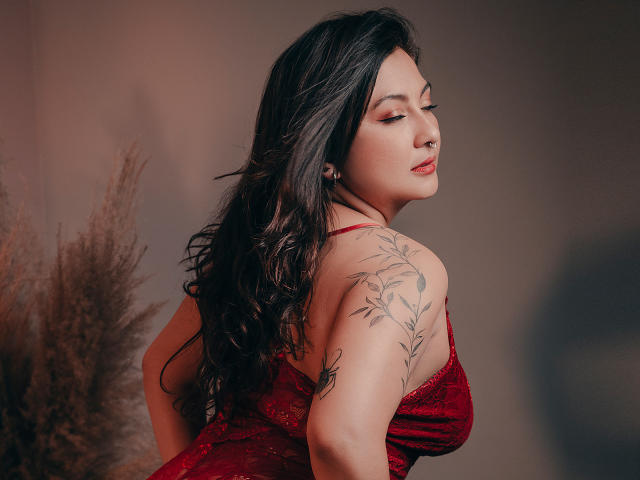 Karlaparadise with long dark hair, red dress, tattoo on right shoulder, eyes closed, red lipstick, soft light, puffy brown grass in background. Boobs visible, curvy, sexy.