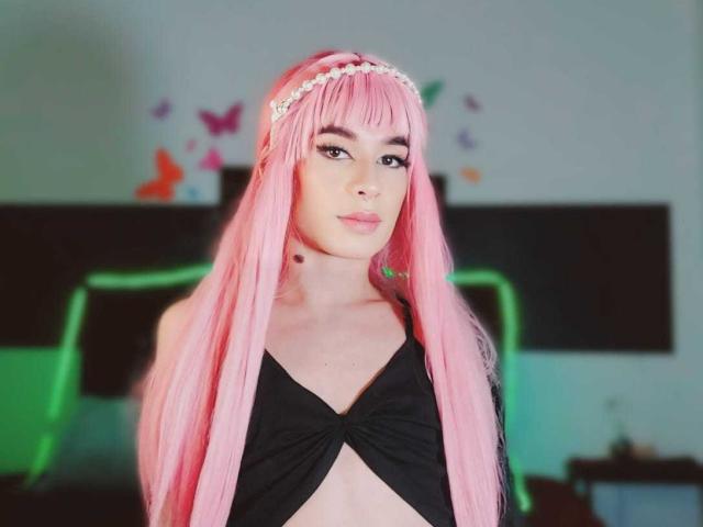 SweetyLy, con i suoi lunghi capelli rosa, indossa una canottiera nera, ha una fascia per capelli di perle e una piccola lentiggine sul collo, e guarda la fotocamera con un leggero sorriso..