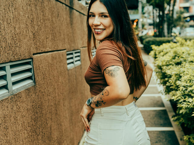 SophiaMarx with long brown hair, brown crop top, white high-waist skirt, smiling, tattooed arm, wristwatch, standing against textured brown wall, looking back, showing her ass.