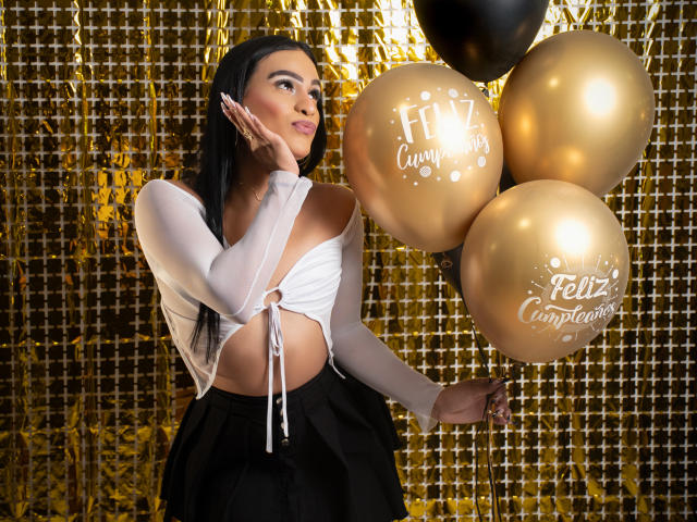 IrinaFerrero with long black hair, white off-shoulder top, black skirt, holding gold balloons, hand on cheek, golden background, sexy, cute, attractive, black balloon, festive, confident.