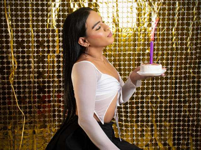 IrinaFerrero with long black hair, white sheer top, black skirt, kissing a white cupcake with purple candle, her eyes closed, holding the cupcake in her right hand.