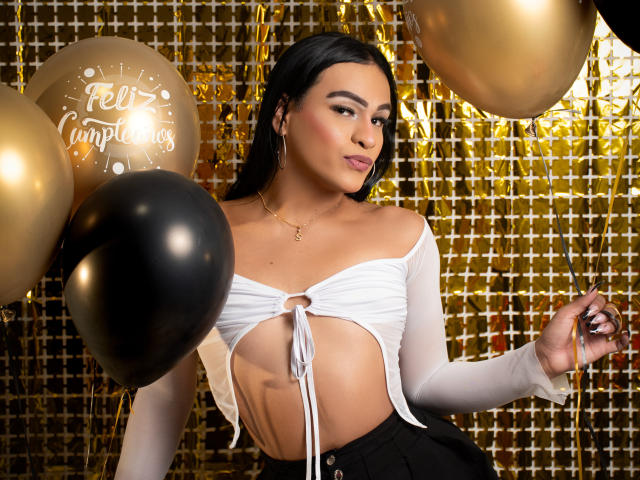 IrinaFerrero with black hair, wearing a white tied-up top showing her tits, holding black and gold balloons, golden background, and a gold necklace. She's looking at the camera.