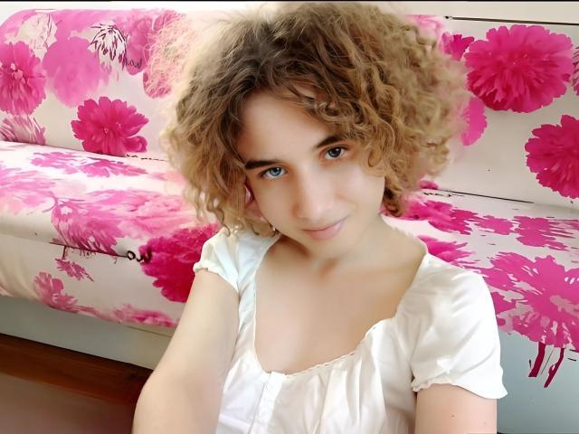 SelenaXCute with curly blonde hair, blue eyes, and white blouse, showing small tits, sitting in front of pink floral sofa, smiling softly.