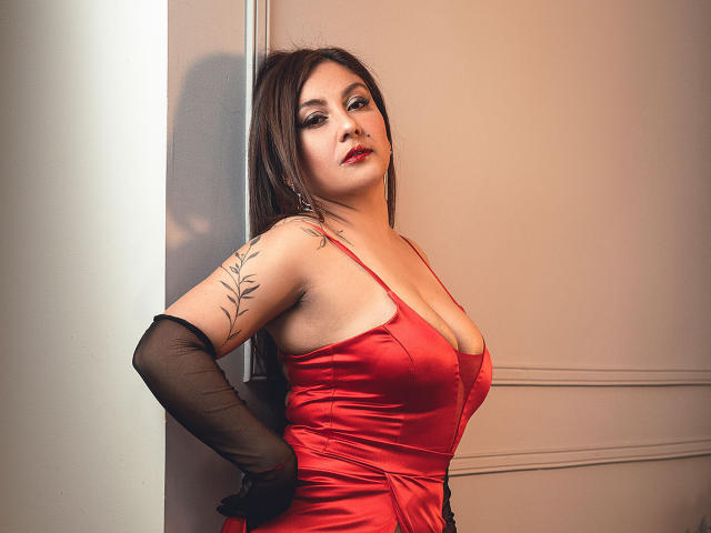 Karlaparadise with big boobs in a red satin dress, black gloves, and a leaf tattoo on her arm, leaning against a wall, looking sexy with red lipstick.