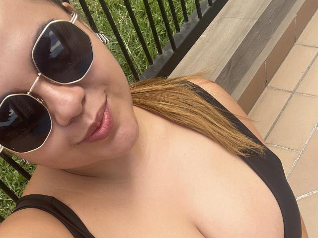 MissSofiia with round sunglasses, black tank top, and pouty lips, showing her tits and light brown ponytail in a selfie. Her skin is tan, and she's smiling slightly.