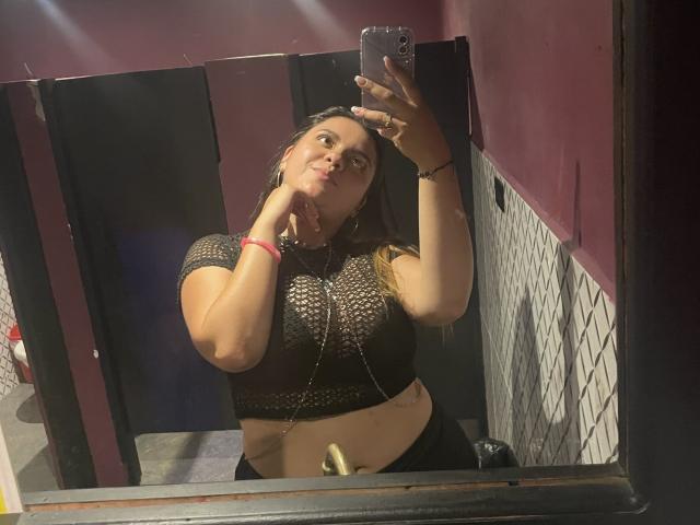 Mirror selfie of MissSofiia with her hand on her chin, wearing a black crochet top, showing her boobs, in a dimly lit bathroom, holding a phone.
