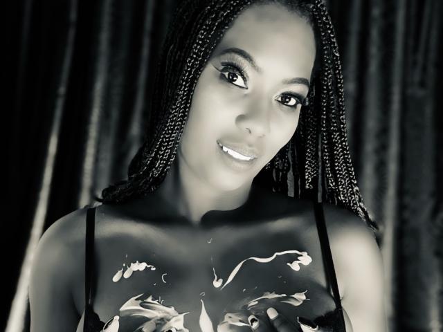 SaraMarks with dark braids, smiling, black tank top, white liquid on chest, black background, her tits visible through the top, looking at camera, smiling with white teeth.